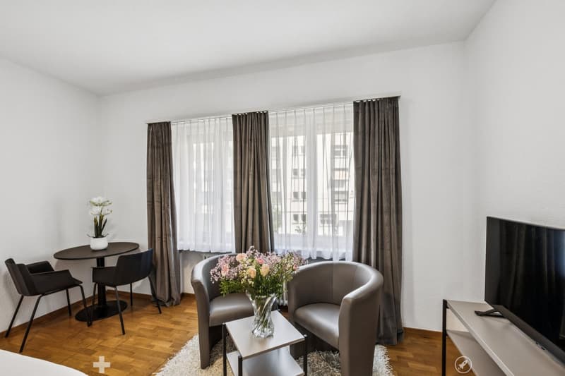 Spacious living room with large windows, hardwood floor, dining table, armchairs, and a vase of flowers on a side table