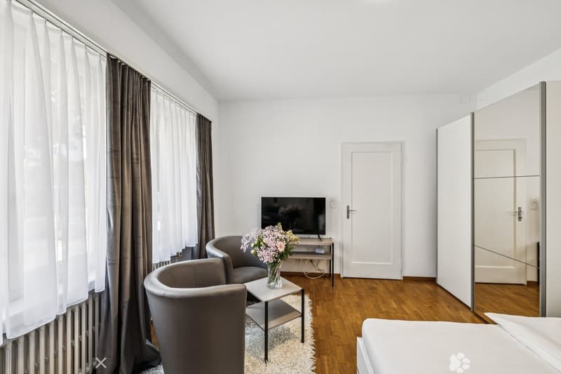 Spacious bedroom with large windows, hardwood floor, gray armchairs, coffee table, and a TV stand. The room has a modern and minimalist design.