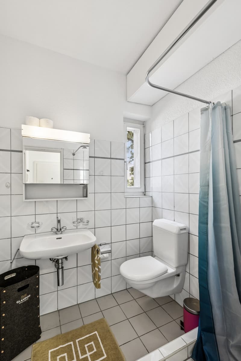 The image shows a bathroom with white tiled walls and floor, a sink, a toilet, and a shower curtain. The bathroom appears to be well-lit with a vanity mirror and lighting fixture above the sink. There is also a small rug on the floor and a storage contain