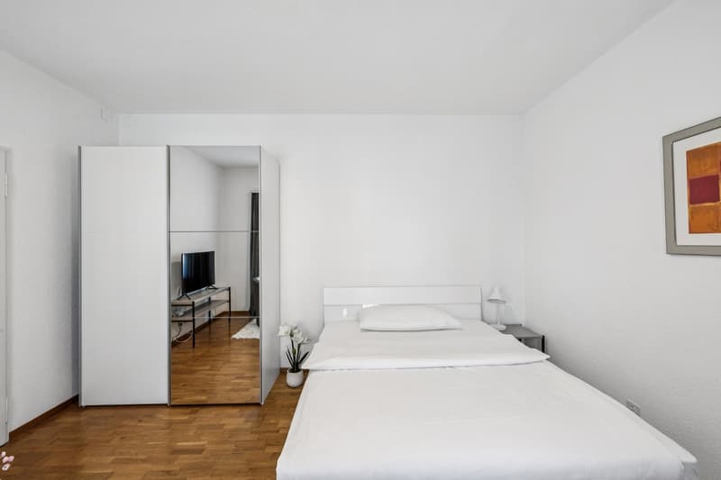 Spacious bedroom with white walls, hardwood flooring, a large bed with white bedding, a mirrored wardrobe, and a TV stand.