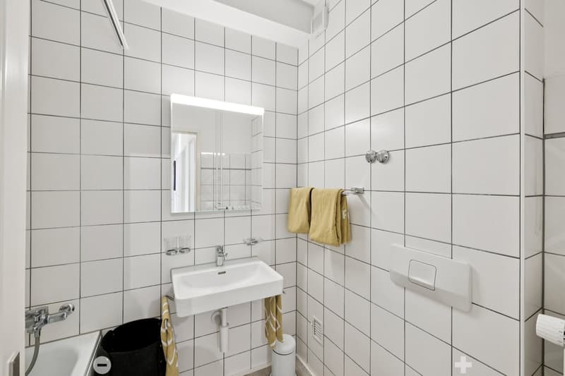 white tiles, small white sink, small mirror, bathtub, waste bin, white toilet, yellow towels