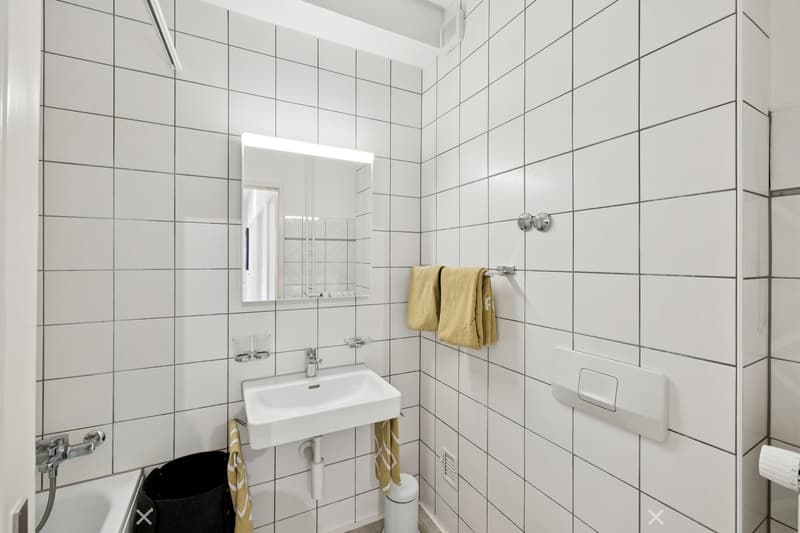 white tiles, small white sink, small mirror, bathtub, waste bin, white toilet, yellow towels