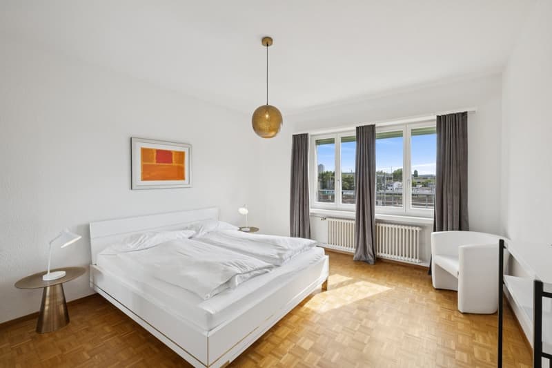 white walls, wooden floor, large window with view, white bed with white bedding, table with white lamp, white radiator, white chair, framed painting on the wall