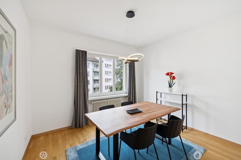 wooden table and chairs, blue rug, white walls, window with view, radiator, shelf with flower