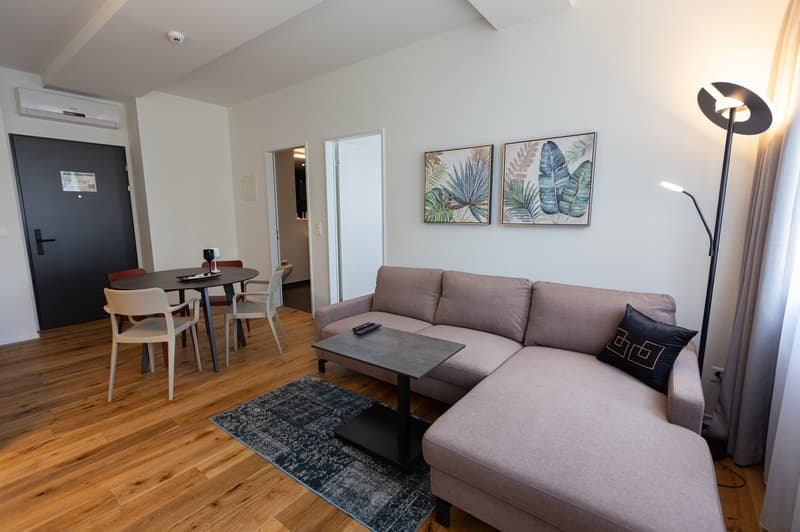 Spacious living room with hardwood floors, gray sofa, coffee table, and dining area with a round table and chairs. The room is decorated with tropical-themed artwork on the walls and has a floor lamp and air conditioning unit. There appears to be a balcon