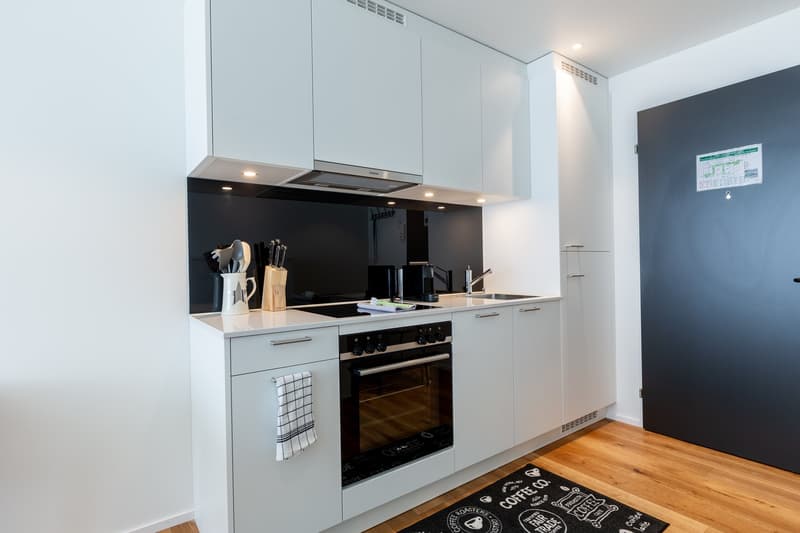 Modern kitchen with white cabinets, black countertop, built-in oven, and dishwasher. The kitchen also has a black backsplash and wooden flooring.