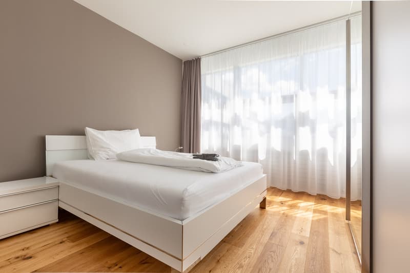 Spacious bedroom with white bed frame, white bedding, and large windows with sheer curtains providing natural light. The room has hardwood flooring and a neutral color scheme.
