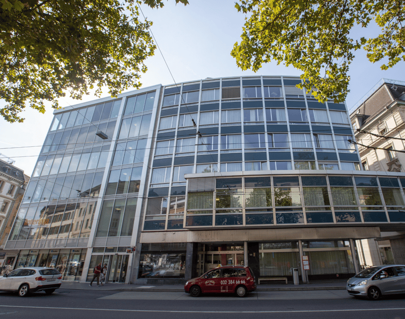 A multi-story commercial building with a glass facade and a modern architectural design. The building has several floors and appears to be used for office or business purposes. There are cars parked in front of the building, indicating it is located in an