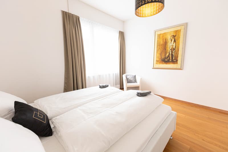 Spacious bedroom with large windows, white bedding, wooden floor, and a framed artwork on the wall. The room also features a pendant light fixture and curtains.