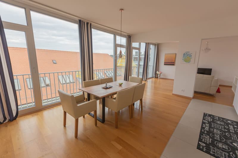 Spacious open-plan living and dining area with large windows and a balcony, hardwood floors, dining table with chairs, and a TV stand