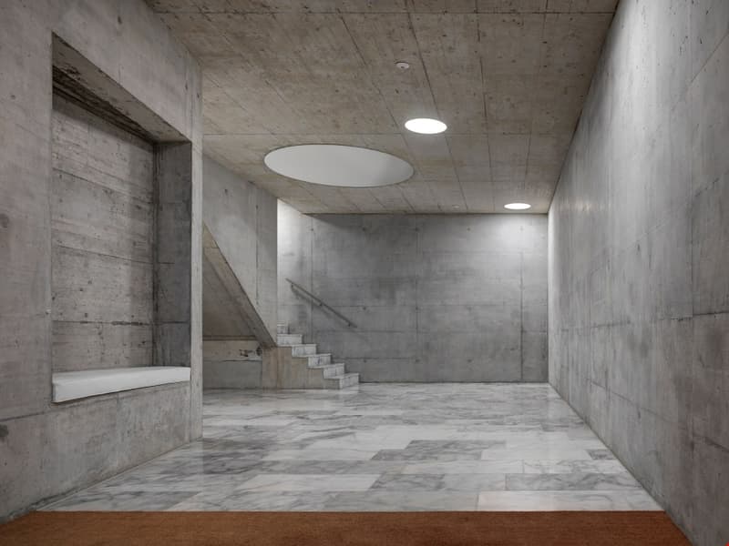 Large concrete interior space with a marble floor, concrete walls, and a staircase leading up. The space is illuminated by circular ceiling lights.
