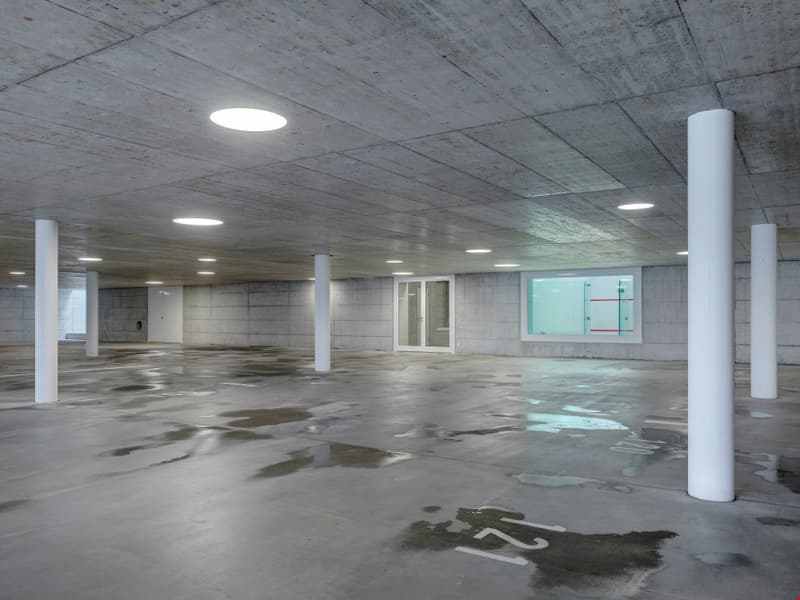 Large open indoor parking garage with concrete floors, columns, and ceiling. The space appears to be empty and dimly lit with recessed lighting.