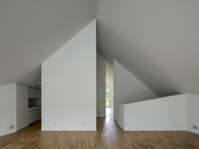 Minimalist attic with wooden floors, white walls, and a small kitchen area. There are slanted ceilings and open-space layout.