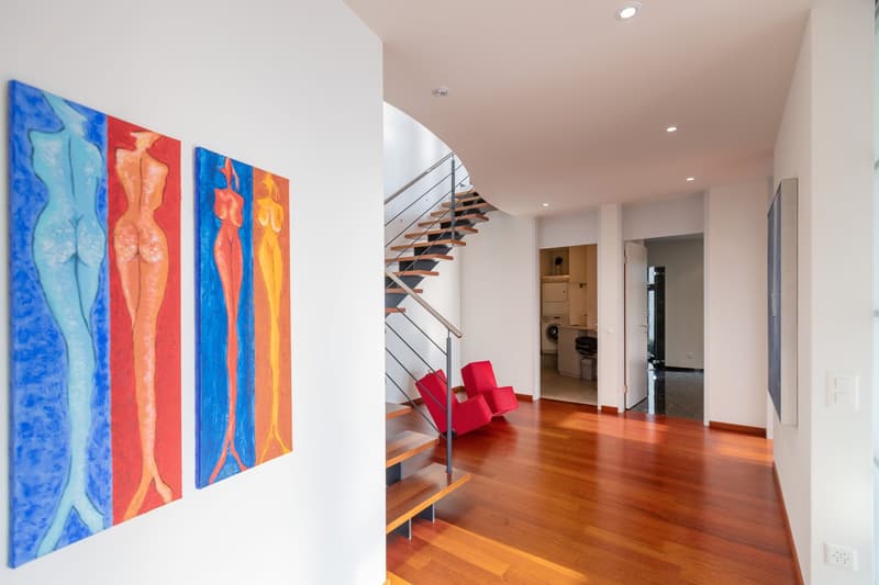 Interior hall with three abstract paintings, wooden floor, staircase, two red chairs.