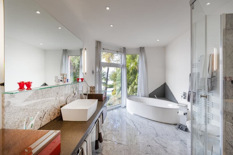 white countertop, large mirror, glass shower, bathtub, white sink, white walls, large windows, gray flooring