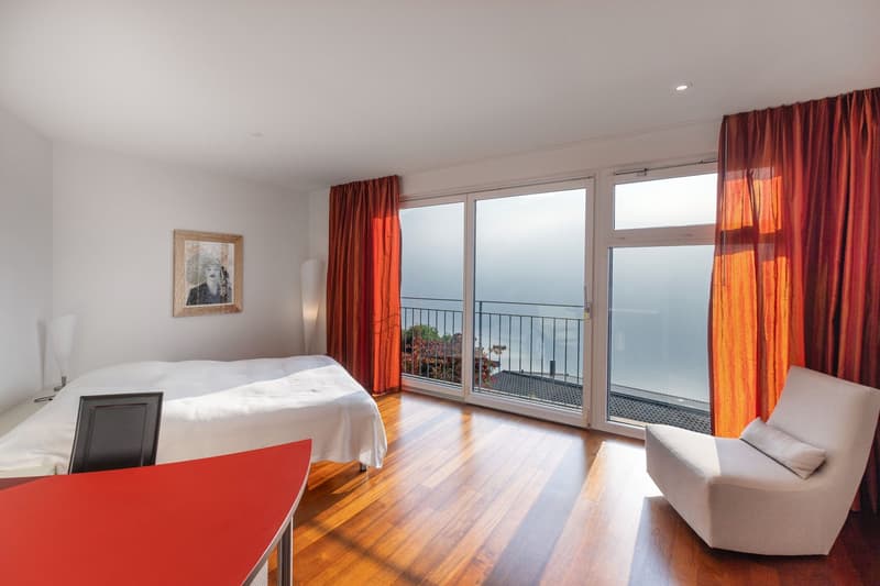 bedroom with wooden floor, glass windows, balcony with sea view, white bed, armchair, desk, painting