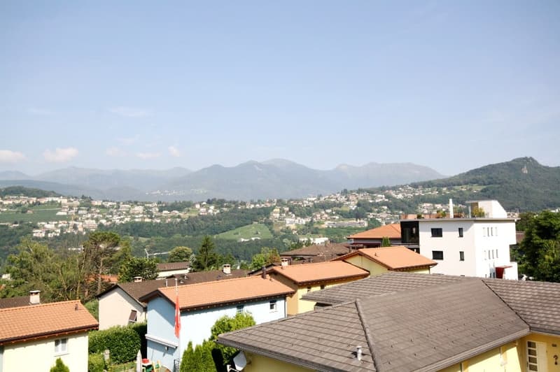 The building appears to be a large home with multiple windows and a sloped roof. A flag is visible near the building, possibly indicating some sort of special occasion or national celebration. The building is situated in a valley surrounded by mountains a