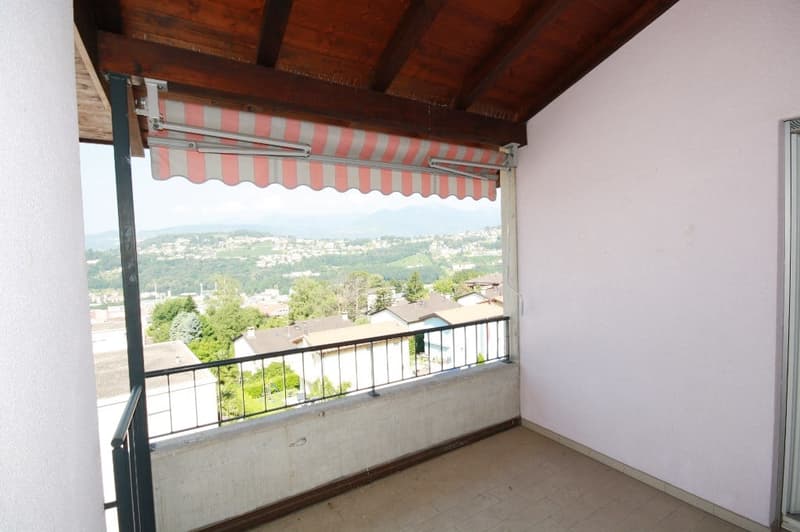 A balcony with a tiled floor, metal railings, wooden ceiling, and a retractable awning. The balcony provides a beautiful view of the cityscape and greenery.