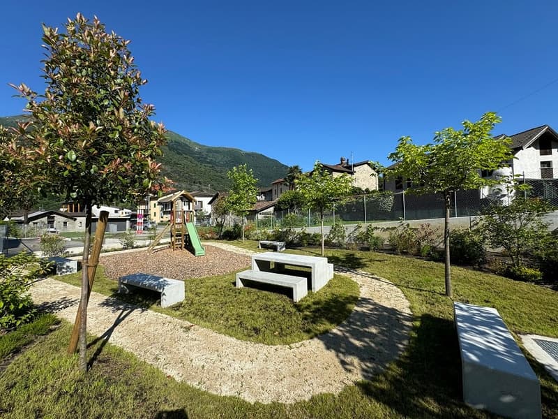 well-manicured lawn, concrete benches, gravel path, trees, playground with slides, houses and mountains in the background