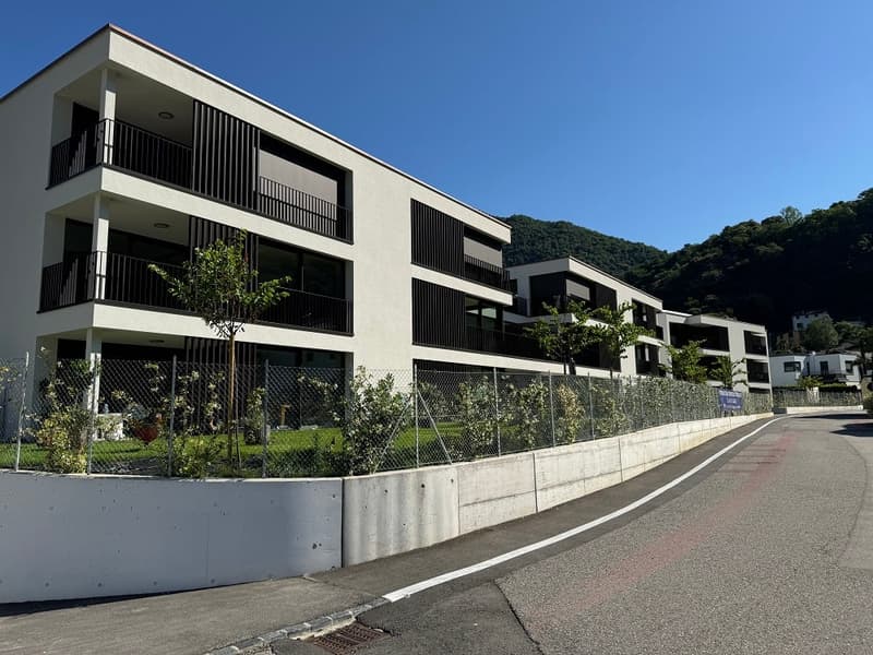 Multi-story apartment building with balconies, concrete and glass facade, surrounded by greenery and a paved walkway