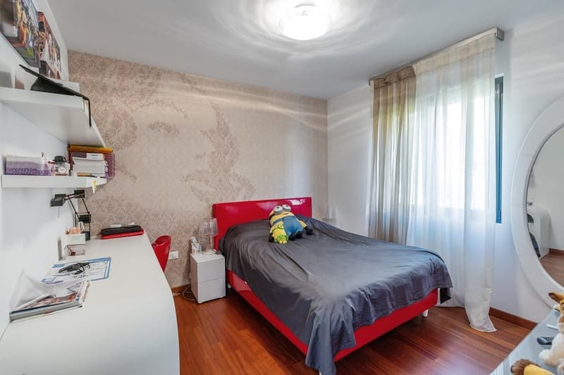 Bedroom with red bed, white desk, white shelves, wooden floor, white walls, white curtains, ceiling lamp, stuffed toys, books, and a mirror.
