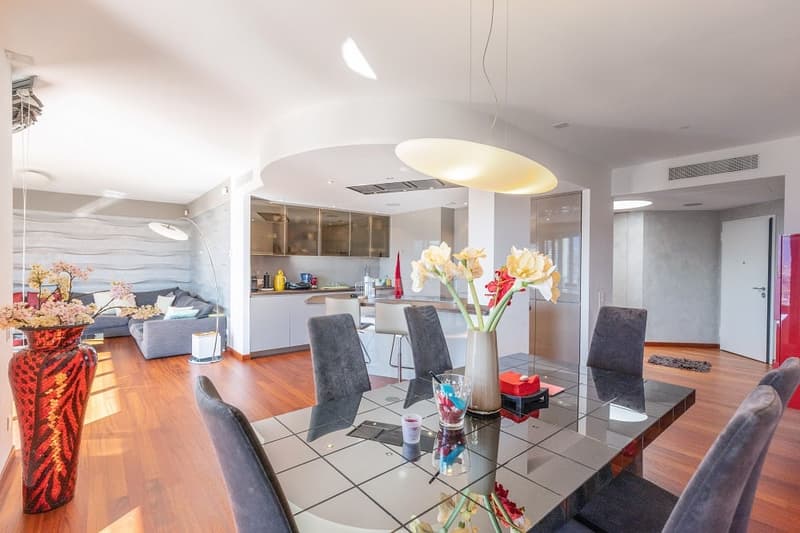 Dining room with a glass table, gray chairs, and a vase with flowers. The room has a modern design with wooden floors and white walls. There is a kitchen area with a countertop and a refrigerator. A couch and a lamp are in the living room area.