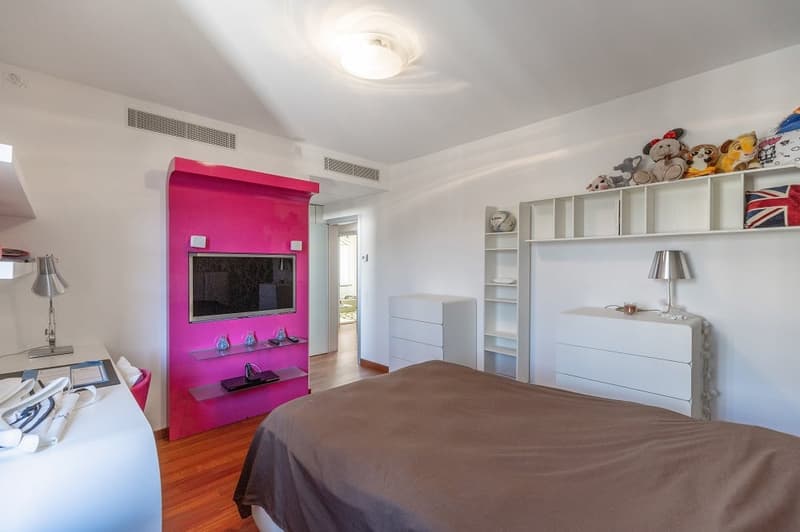 Bedroom with pink accent wall, flat screen TV, wooden floor, white furniture, shelves, lamp, bed, desk, lamp, and toys