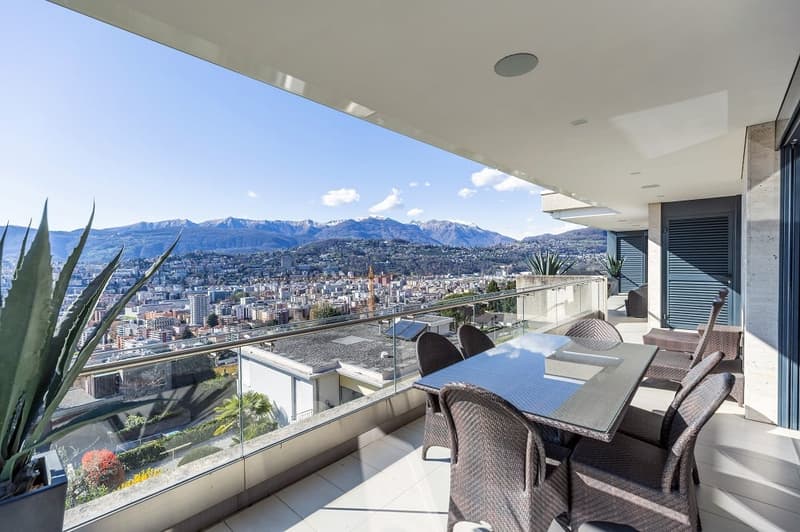 Balcony with table and chairs, glass railing, plants, city view, mountains