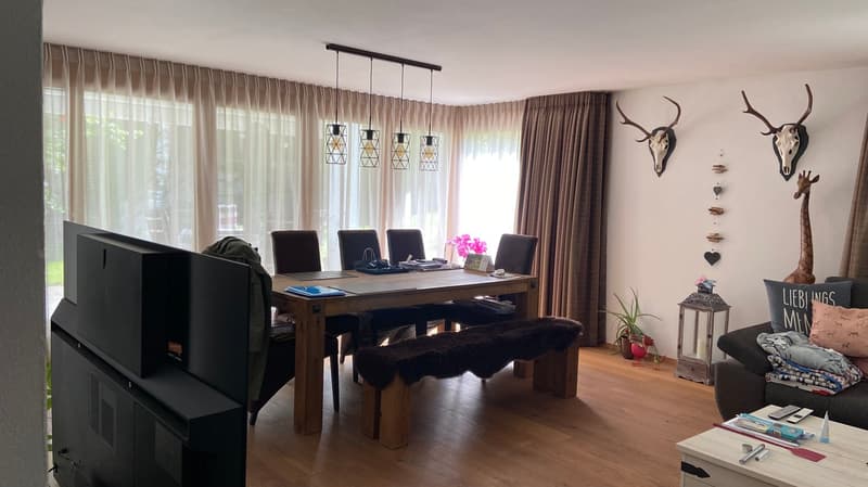 wooden floor, white walls, large windows with curtains, dining table with 6 chairs, bench, couch with pillows, television, wooden table with potted plants and pink flowers, pendant lights, wall decorations