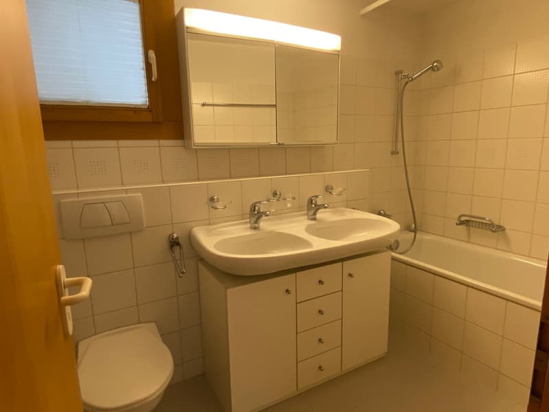 Bathroom with a double sink, white tiles, mirror above the sink, a bathtub, and a toilet. Double sink with two faucets.