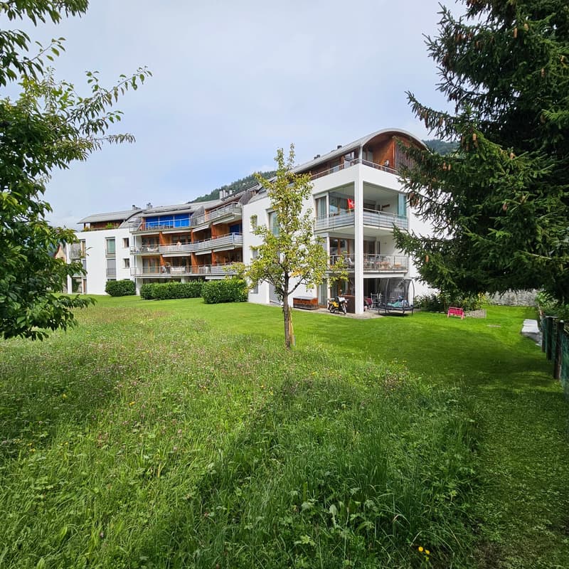 modern white apartment building with multiple balconies, grassy lawn, trees, mountain backdrop, outdoor storage box, small red bench, small trampoline in yard, parked motorcycle