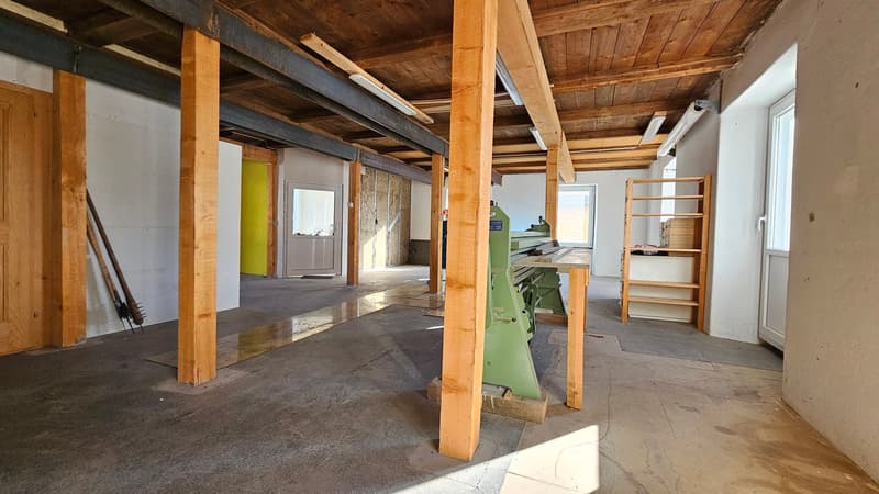 An empty room with wooden pillars, wooden beams on the ceiling, wooden and metal shelves, a window, and a door. The floor is made of concrete.