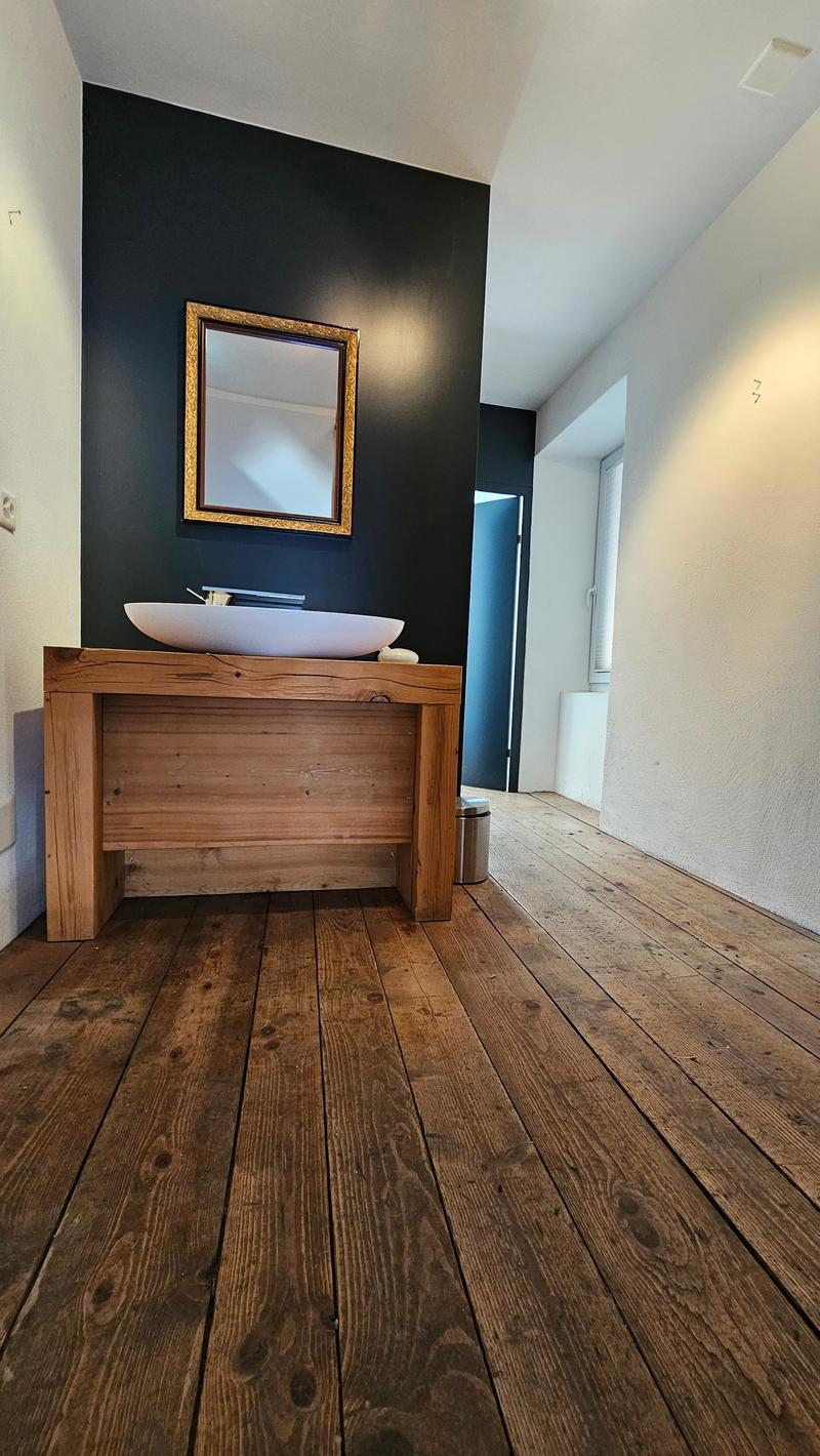 Bathroom with wooden plank flooring, wooden sink, black painted wall, mirror mounted on wall, power outlet on the wall, trash can under the sink.