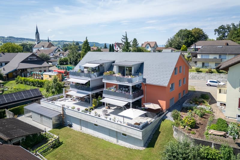 3-story apartment building, orange and gray exterior, multiple balconies with tables and umbrellas, landscaped garden with plants and flower beds, solar panels on the roof, parked car, surrounded by other houses and a church.
