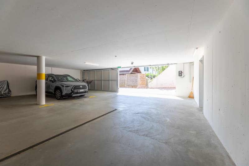 an indoor parking area, gray SUV parked, white walls, gray floor, yellow markings on the floor, an open garage door, ceiling lights, broom near the wall, a wooden shed in the distance