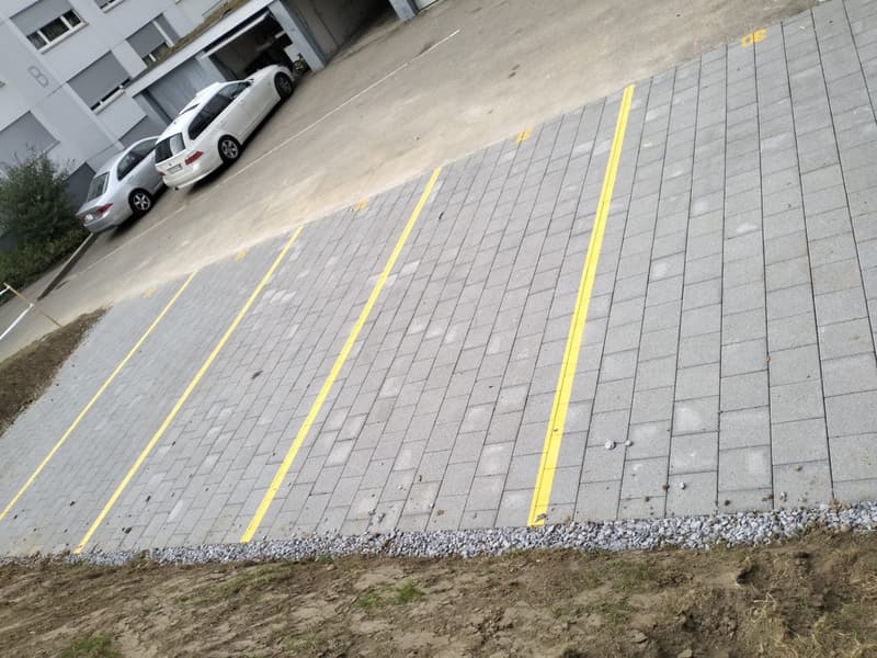 Newly paved parking lot with gray bricks and yellow demarcation lines. Two cars are parked next to the lot.