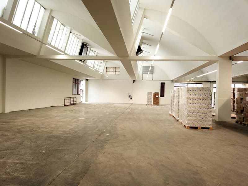 Empty warehouse with high ceilings, large windows, concrete floor, and storage boxes.