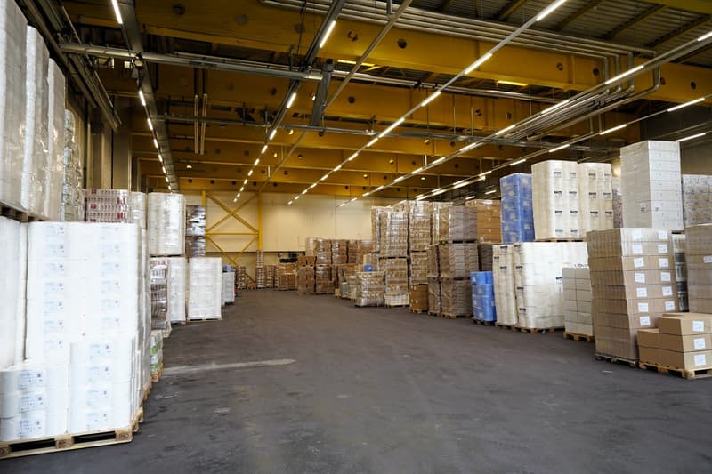 Industrial warehouse with white and blue stacked boxes on pallets, yellow metal beams, lights, and an open space for storage