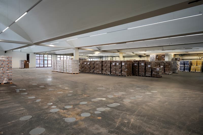 Warehouse with many pallets of goods, long ceiling with recessed lighting, glass windows on the left, grey concrete floor, large empty space