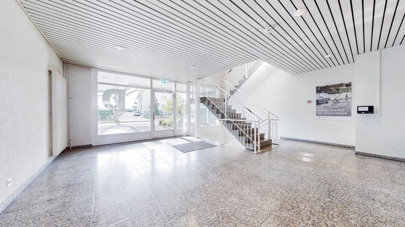 A building exterior with a clean entrance, grey carpet at the stairs, and glass doors leading to the outside. Modern decor, white walls, and ample lighting.