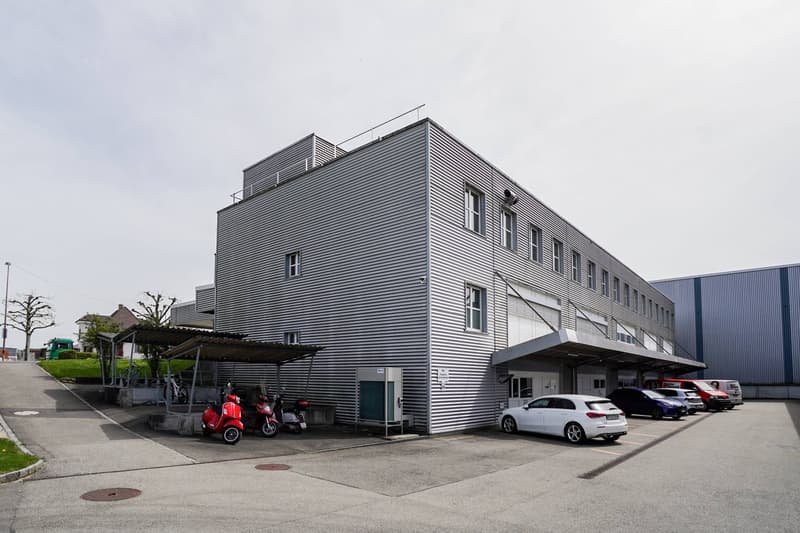 modern architecture, gray exterior, large windows, multiple vehicles parked, metal cover at entrance, flat rooftop