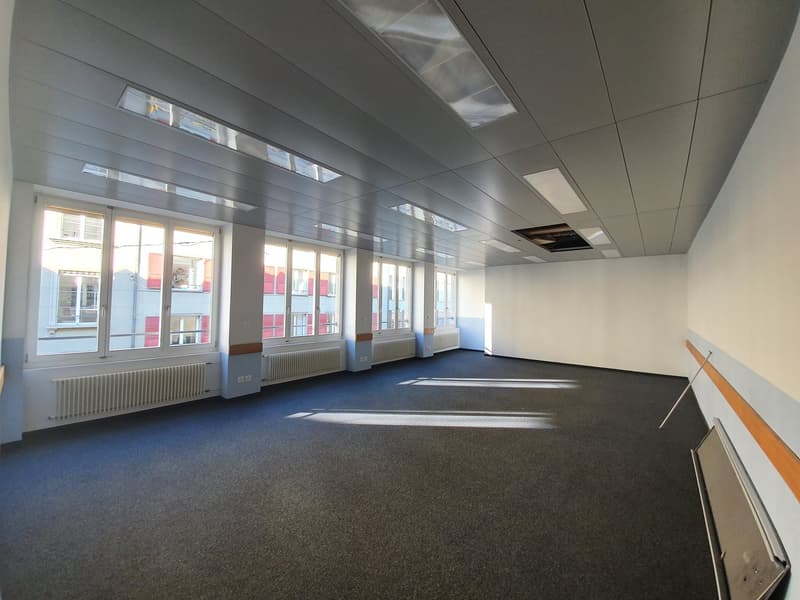 The image shows a large, open office space with gray walls, a tiled ceiling, and large windows along one side. The floor is covered in a dark gray carpet. The space appears to be empty and ready for use as an office or commercial space.