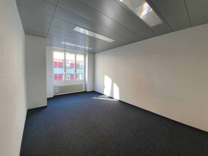 The image shows a spacious office room with a gray ceiling, white walls, and a large window overlooking a city. The room has a blue carpeted floor and a radiator on the wall. The room appears to be empty and ready for use.