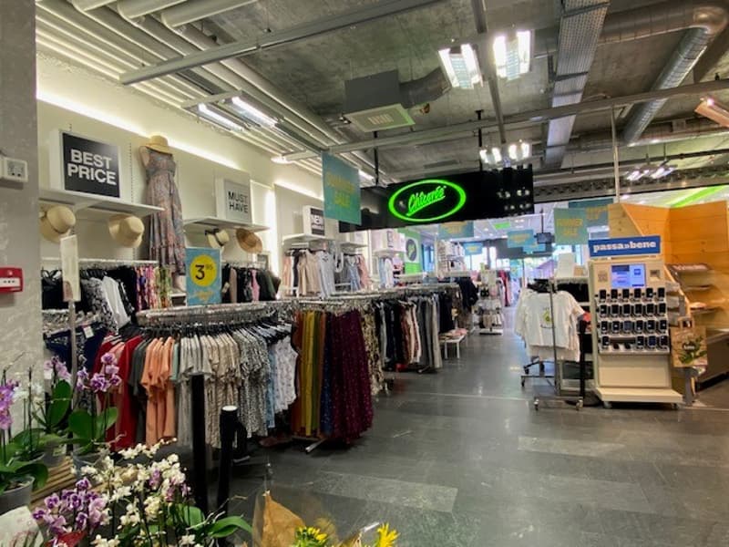 Rows of clothing hangers, clothing displays, clothing store signs, white ceiling lights, shopping cart, flower arrangements, mannequin heads on white shelves