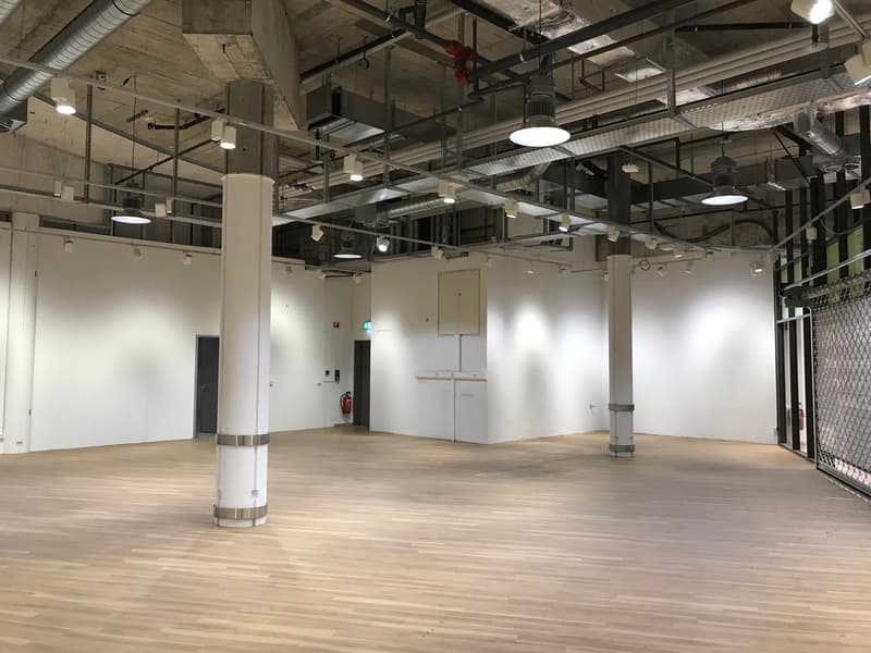empty room with white walls, wooden floor, suspended lamps, fire extinguisher, columns, and emergency exit sign