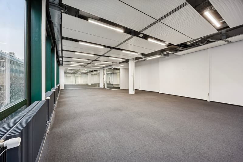 Empty room with large windows, gray floor, and white walls. The ceiling has a grid structure with recessed lights. There are radiators on the left side, and a white ceiling above.