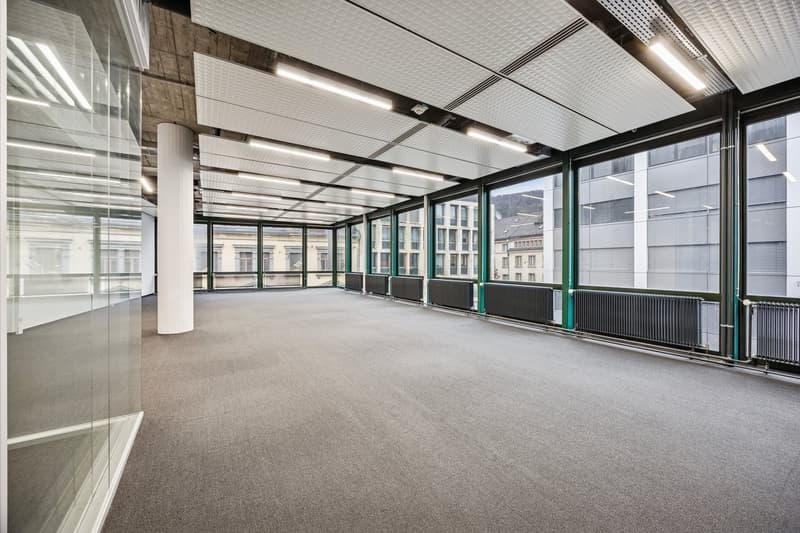 Empty office with large glass windows, carpeted floor, modern ceiling and large pillars.
