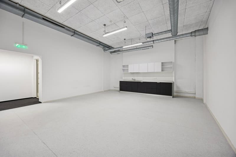 An empty, plain room with white walls, ceiling, and floor, featuring a kitchen area with cabinets and a sink.