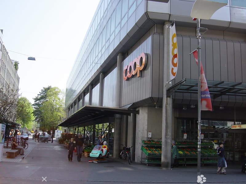 Gray building with the words 'COOP' on it, has awning, bicycles parked in front, person sitting on bench, several people walking on the street, fruit baskets on the side, and street lights