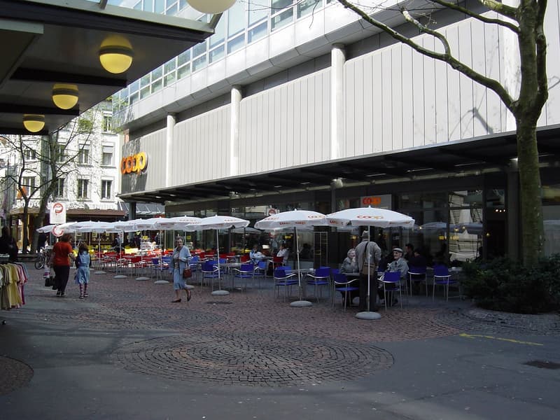 An outdoor seating area with tables and chairs under white umbrellas at a restaurant, surrounded by plants and trees with a brick floor, beside a large building with signage and an open entrance